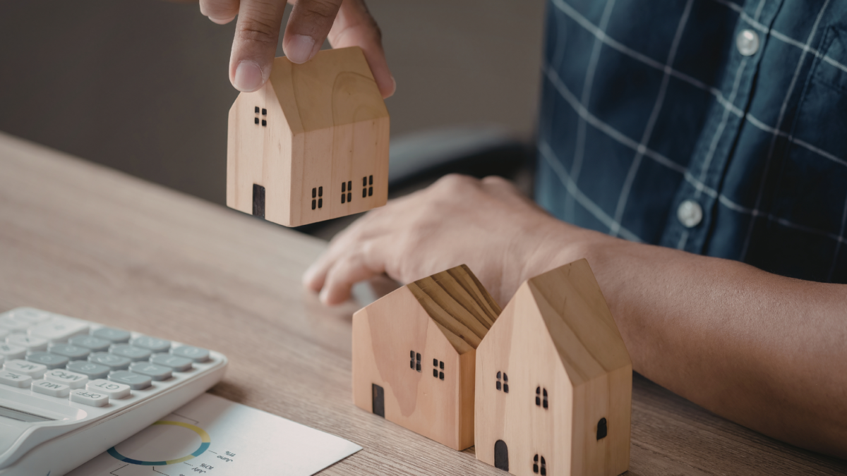 Hand placing a small wooden house model next to two other miniature wooden houses on a desk, symbolizing property or real estate planning.