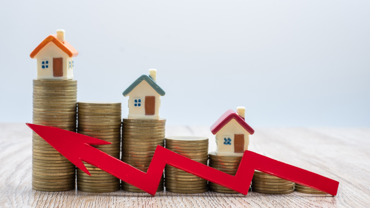 Stacked coins with miniature houses and a red downward arrow representing declining property values or a housing market downturn.