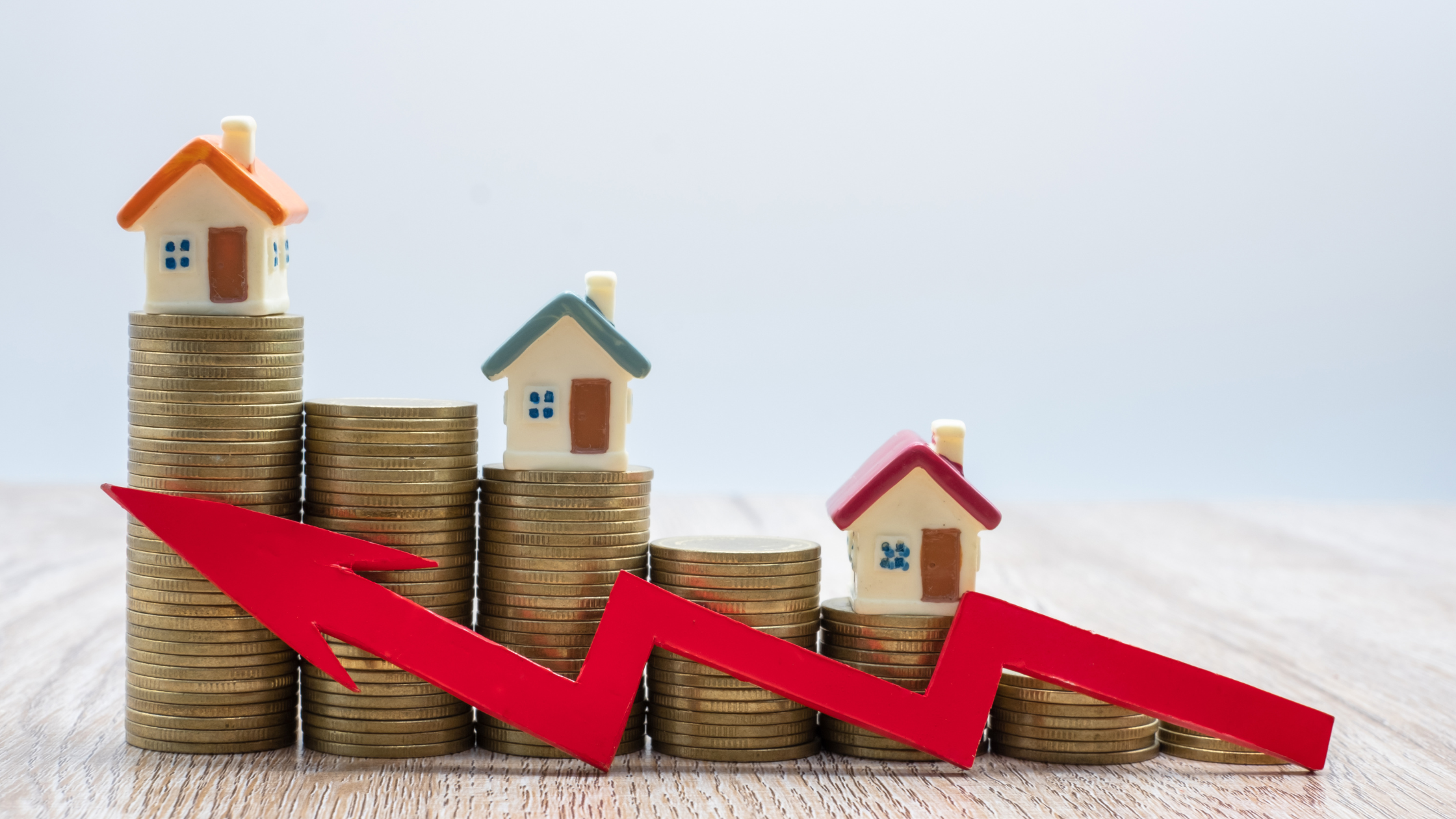 Stacked coins with miniature houses and a red downward arrow representing declining property values or a housing market downturn.