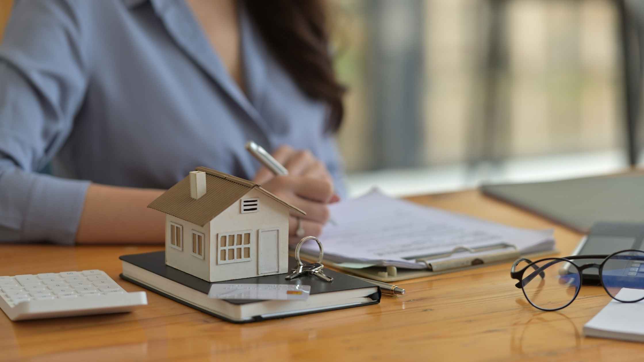 Property investment professional reviewing documents beside a model house, calculator and keys on a desk.